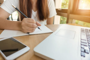 Eine Frau sitzt an einem Schreibtisch und schreibt mit einem Bleistift in ein Notizheft.
